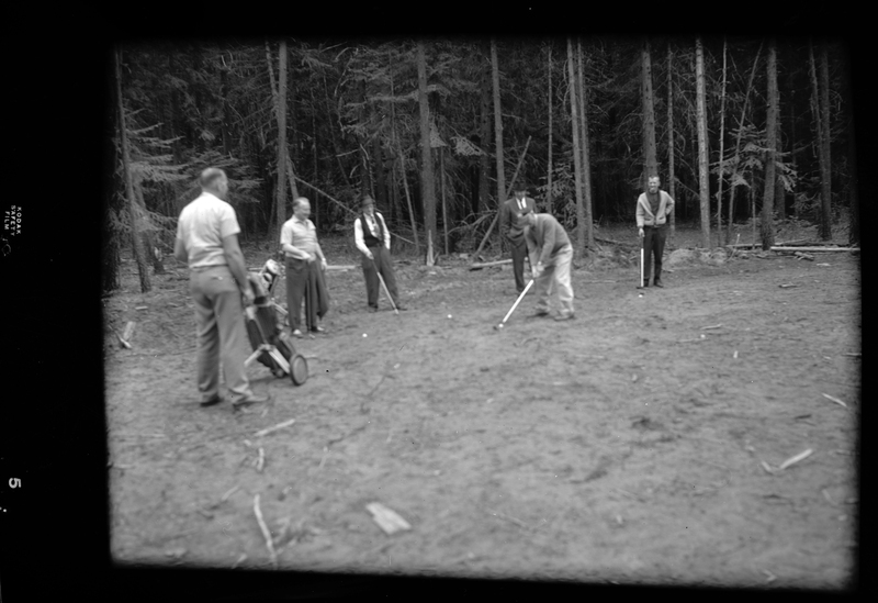 Six men can be seen golfing at Big Creek. There are trees surround them all and one man is actively about to swing.
