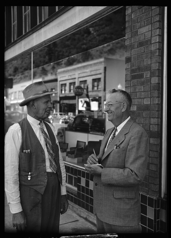 Mayor Betts (left) is talking to Steve Eddins (right). They are standing outside and the reflection of a street with buildings can be seen in the reflection of the window behind them.