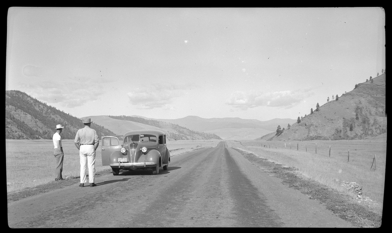 Two men are standing by a car, which is pulled off to the side of the road, and looking down the long stretch of road behind the car. The road appears to be unpaved, and hills are visible in the background.
