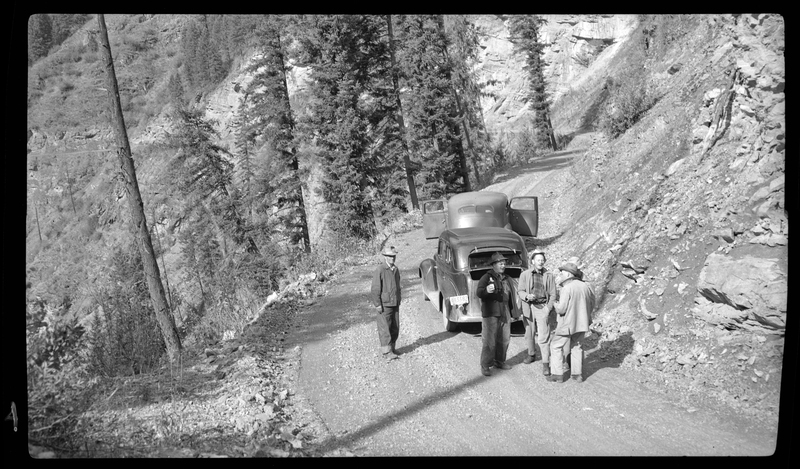 Several men stand next to two cars, holding drinks and talking. They are parked on the side of an unpaved road in what is likely a mountainous area.