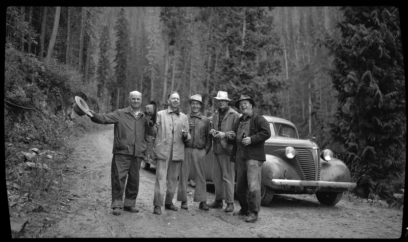 Several men stand in front of a car, holding drinks, and some are holding out their hats. The car is parked on an unpaved road and there are trees surrounding them.