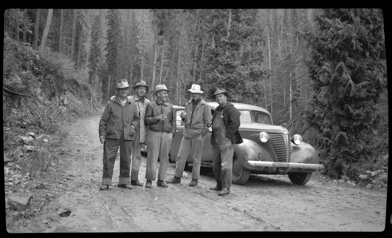 Several men stand in front of a car holding drinks. The car is parked on an unpaved road and there are trees surrounding them.