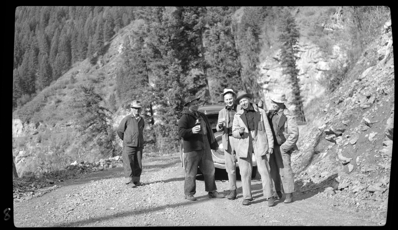 Several men stand next to two cars, holding drinks and talking. They are parked on the side of an unpaved road in what is likely a mountainous area.