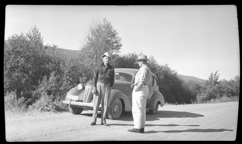 Two men stand by a car that is pulled off to the side of the road.