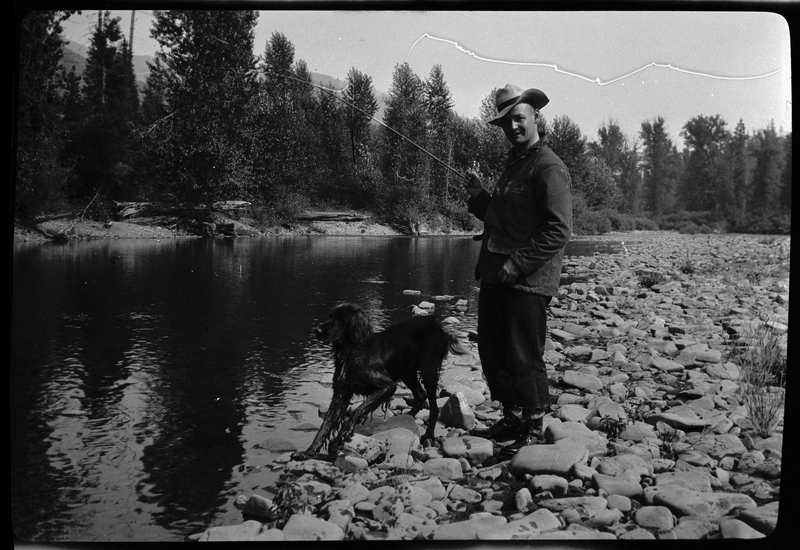 An unidentified man standing on a rocky ledge next to a river holding a fishing pole. There is a wet dog standing between him and the water, and there are trees in the background.