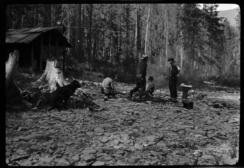 Four unidentified men are lighting a fire on the rocky ground near a cabin. There are trees in the background, and a dog is running over to the men.