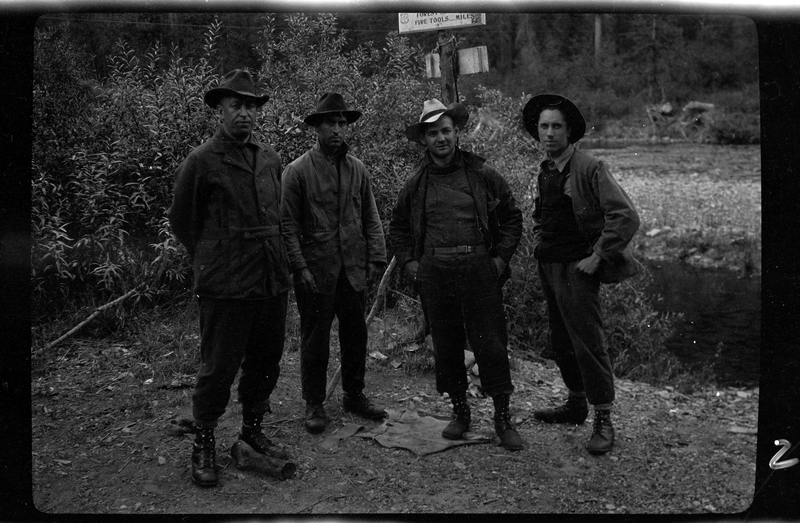 Four unidentified men stand together for a picture. There are trees, a sign, and a river in the background.