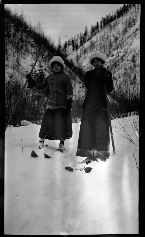 Two unidentified women wearing skis in the snow. They are wearing coats, gloves, and hats, and appear to be digging.