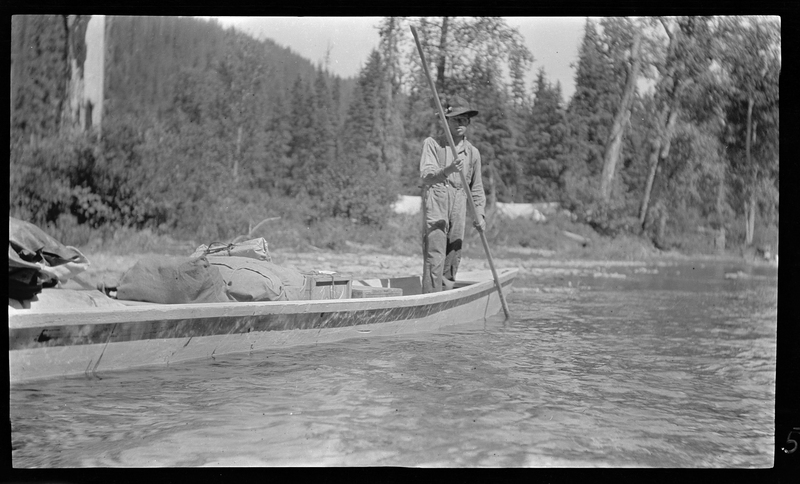 An unidentified man is standing in a canoe and appears to be paddling. There are trees visible in the background.