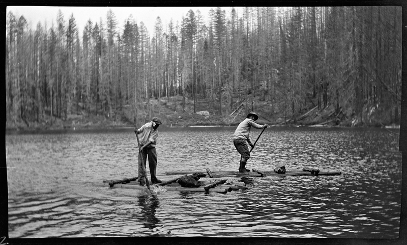 Two unidentified men standing on makeshift rafts paddling in the river. Trees can be see in the background.