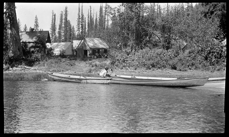 An unidentified man is on the river bank between two canoes. He appears to be loading bags into the boat. There are trees and buildings in the background.