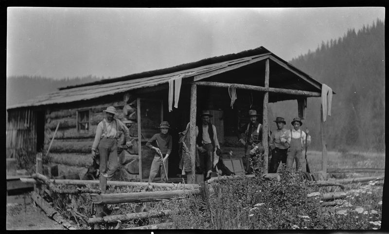 A group of unidentified men standing on the porch of a cabin with a dog. Some of the men are holding guns.
