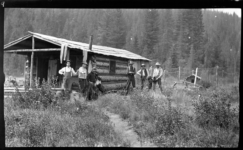 A group of unidentified men standing outside of a cabin with a dog. Some of the men are holding guns.