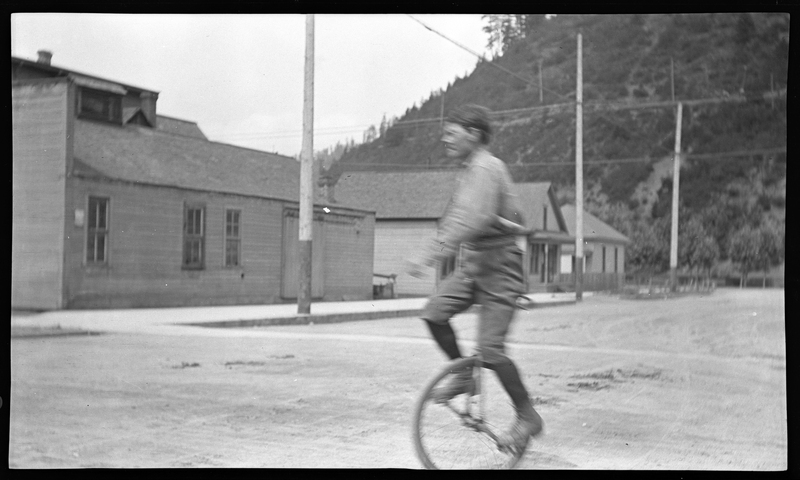 Photo of a man riding a unicycle on a street. The man is blurry from movement. There are buildings and trees in the background.