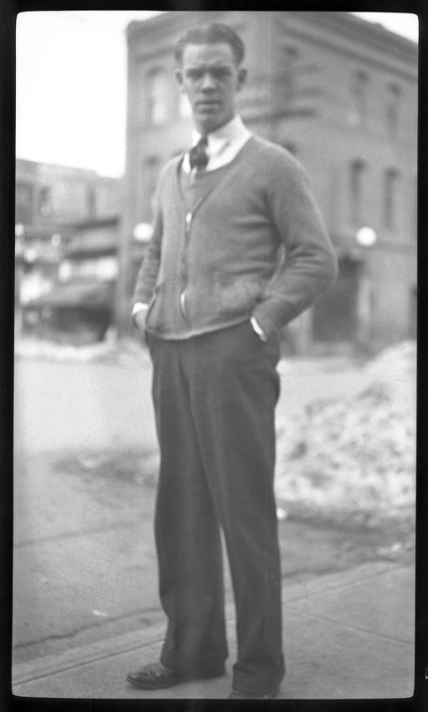Photo of an unidentified man standing on the sidewalk with his hands in his pants. The photo is slightly blurry, but there appears to be a pile of snow behind him, as well as some buildings.