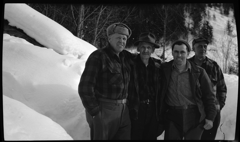 Four unidentified men stand together surrounded by snow. They are all dressed to stay warm and they are all looking at the camera.