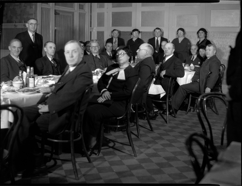 Group of finely dressed men and women (mostly men) sitting or standing around a table. Most of the people in the photo are looking at the camera.
