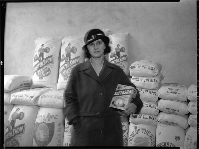 A woman at Stone's Store holding a box of Softasilk flour. There are bags of flour from varying brands stacked up behind her.