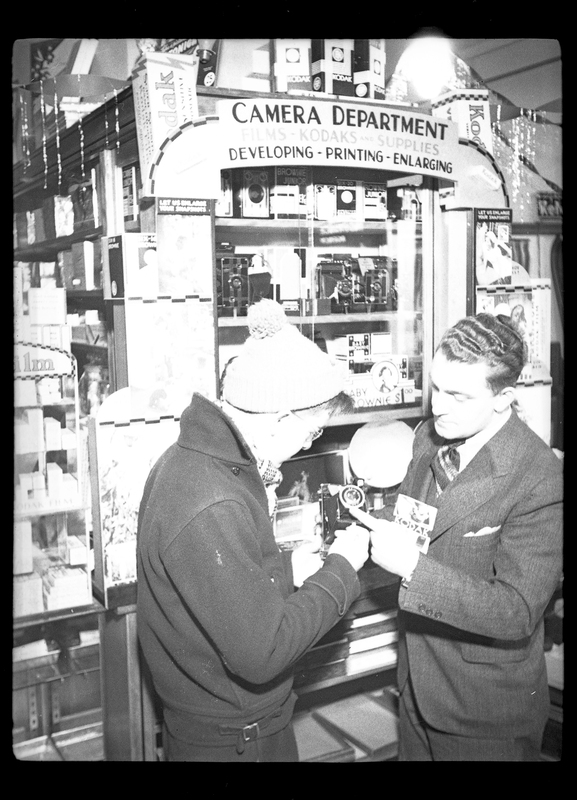 Photo of two people looking at a camera in front of the "camera department" at a store. One man is pointing out something on the camera to the other.