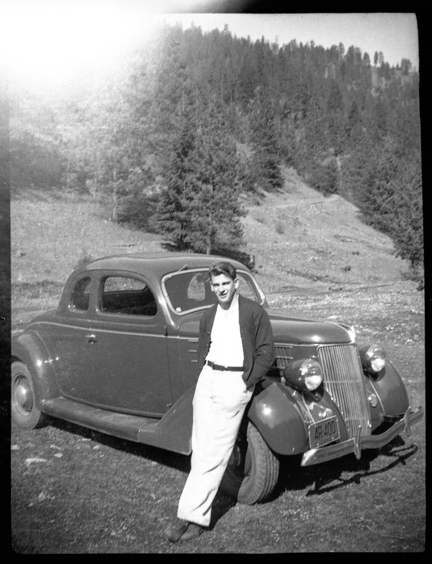 Photo of an unidentified man leaning against a car. There are trees in the background and the license plate on the car reads "4R 400."