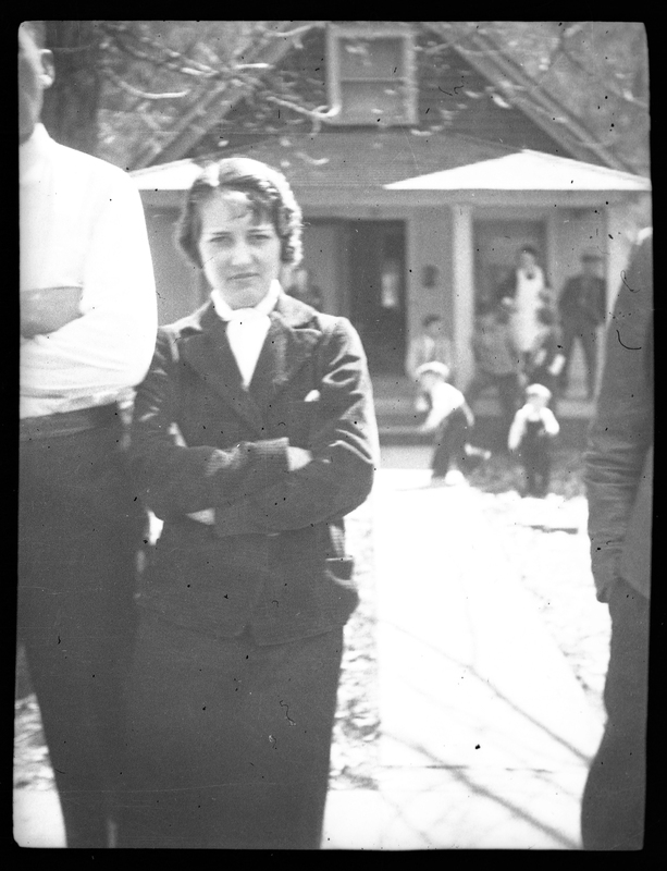 Photo of a woman standing with her arms folded over her chest. There is a person standing next to her that is mostly out of frame, and there are children playing on the porch of a house in the background.