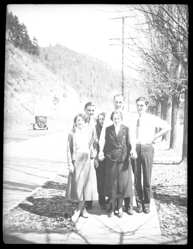 A group of people stand huddled together on the sidewalk, posing for the photo. The photo is overexposed, but a car can be made out in the background, as well as some trees.