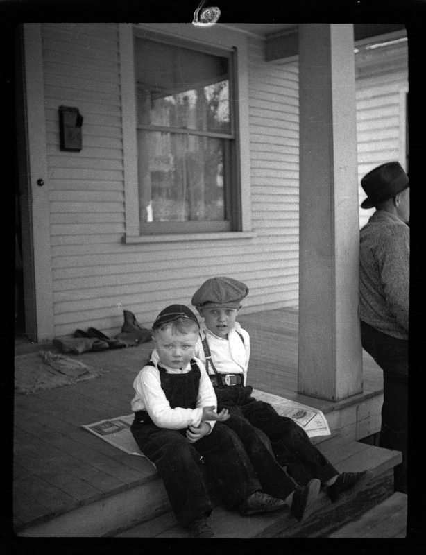 Two children sitting on the top step of a porch of a house. They are wearing similar outfits, and a man can be seen that is almost out of frame.