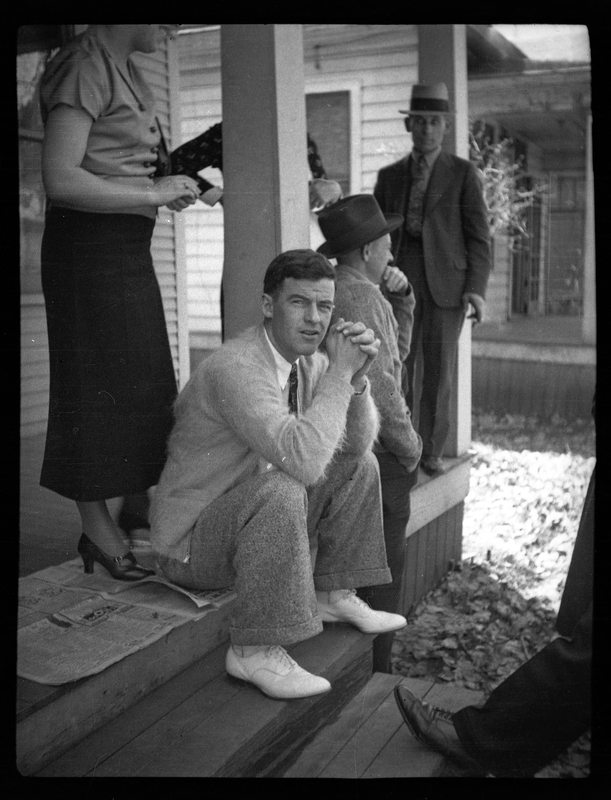 A small group of people on the porch of a house. The main subject of the photo is an unidentified man who is sitting on the top step, looking at the camera directly.