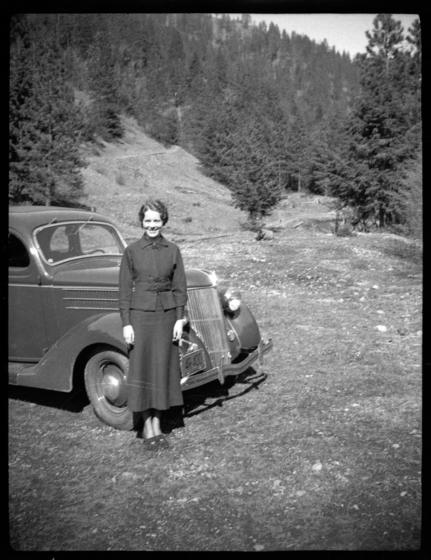 An unidentified woman standing next to a car, with trees and hills visible in the background. The license plate on the car reads "4R 400."