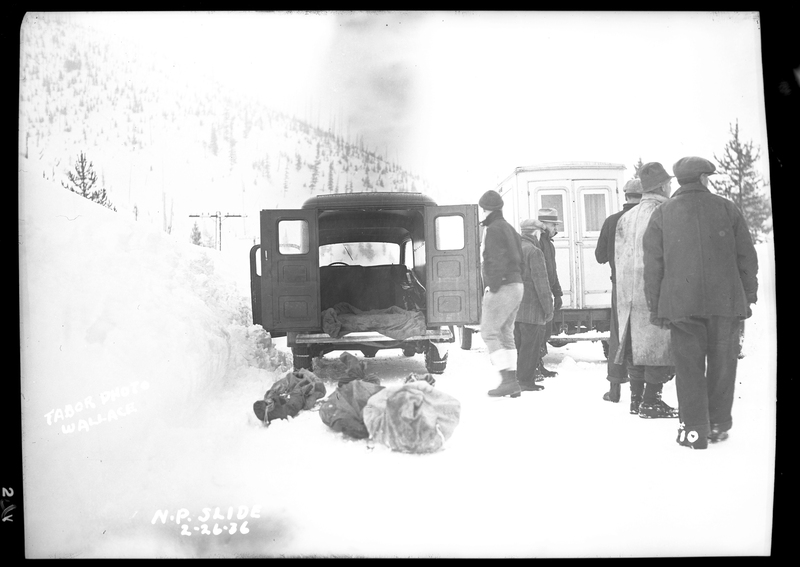 People standing in the snow near two cars, one with the back doors open, and three body bags on the ground in front of it. An avalanche hit a train on the Northern Pacific Railroad. Three people died.
