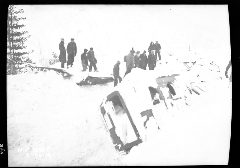 A group of people standing in the snow looking at the wreckage of a train after an avalanche hit it on the Northern Pacific Railroad. Three people died.