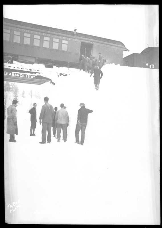 A group of people are climbing up a pile of snow to examine the inside of a train car after it was hit by an avalanche on the Northern Pacific Railroad. There is also a group of people seen in the foreground talking to each other. Three people died.