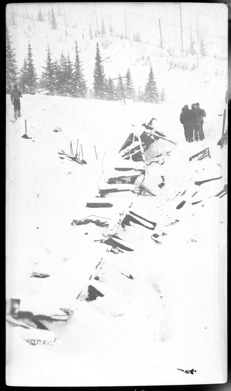 Train wreckage covered in snow after it was hit by an avalanche on the Northern Pacific Railroad. A handful of people can be seen standing next to the wreckage. Photos say that three people died.