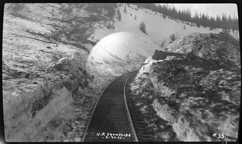 Photo of a set of railroad tracks that appear to have been dug clean of snow. There are walls of snow packed up on either side of the tracks, which curve behind a pile in the picture. The trees on the left side of the photo are knocked over from the avalanche, which other photos of the accident say that three people died after the train was hit on the Northern Pacific Railroad.