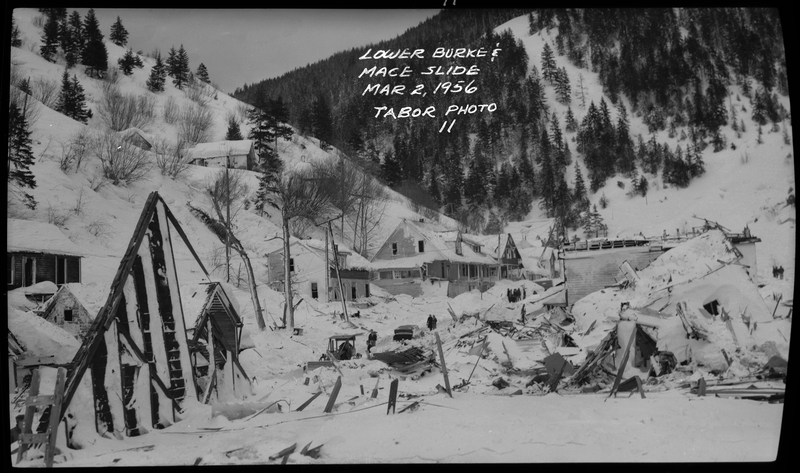 Photo of the damage of sustained by several buildings after an avalanche occurred in lower Burke and Mace, Idaho. The building in the foreground is completely destroyed, and the buildings in the background are in varying levels of destruction. People and snow plows can also be seen in the background.