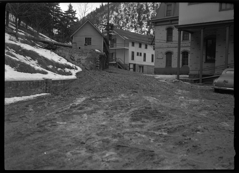 Photo of a small landslide in a populated area with buildings. There is a building, likely a house, in the background, that appears to be damaged from the landslide, but the surrounding buildings appear to be fine. 