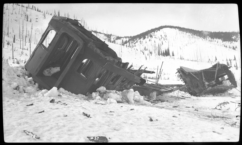 Photo of a damaged train coach that appears to be destroyed as a result of a landslide or avalanche. There is snow on the ground surrounding the wreckage.