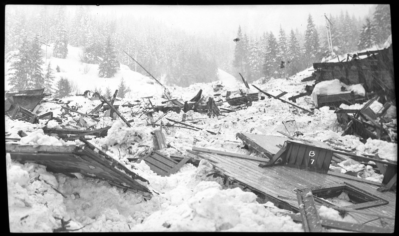 Photo of the damage caused by an avalanche in Burke, Idaho. The damage appears to have been done to buildings, and the area is covered in debris and snow.