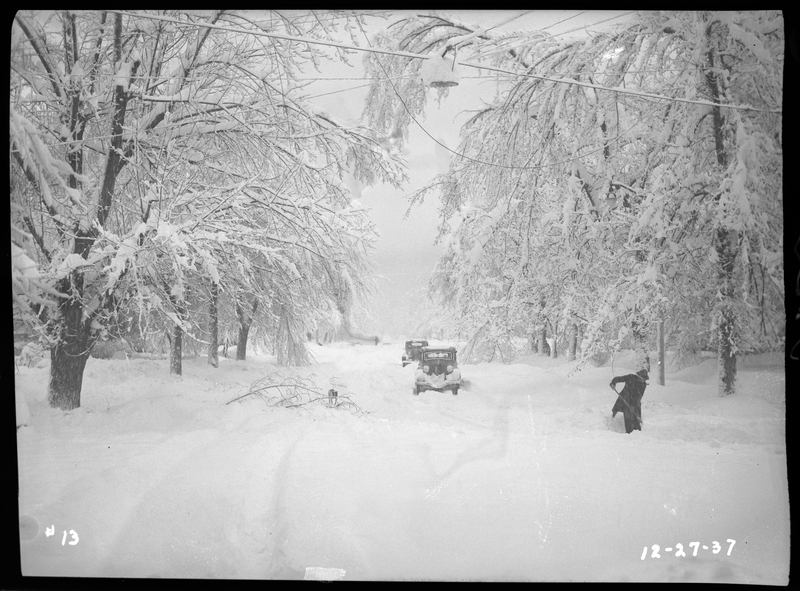 An unidentified person is seen attempting to shovel several inches of snow in Wallace, Idaho during a snowstorm. The bare trees that line the street are covered in snow, as are the two cars on the road. The windshields of the cars are free from snow, suggesting that they are being driven.