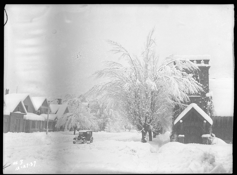 Photo of a car driving down a plowed road during a snowstorm in Wallace, Idaho. The trees and buildings that are visible are covered in several inches of snow, and it is actively snowing as the picture was taken.