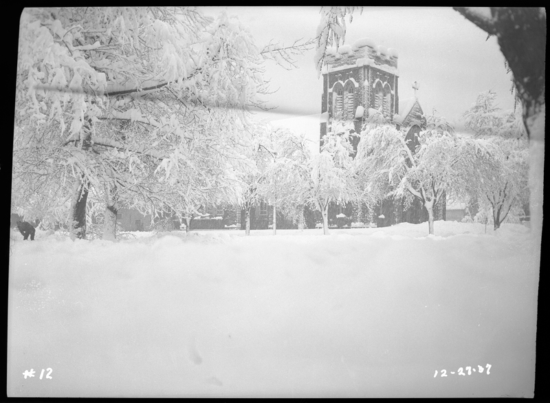 Photo of a church during a snowstorm in Wallace, Idaho. The grounds that surround the church, as well as the trees, are covered in a heavy layer of snow, and it is actively snowing as the photo was taken.