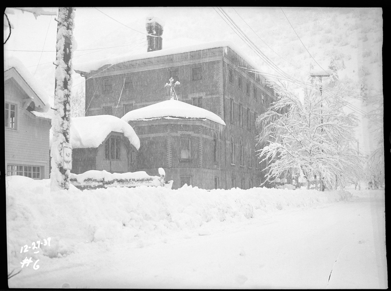 Photo of several buildings in Wallace, Idaho including Our Lady of Louordes Academy during an active snowstorm. The roofs of the buildings, as well as the trees and street are covered in several inches of snow.