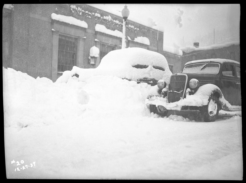 Two cars parked outside of the Wallace United States Post Office. One car is covered in several inches of snow, as well as blocked in from the road behind plowed. The car next to it is more free from snow, suggesting it had been driven recently. It is actively snowing in the photo.