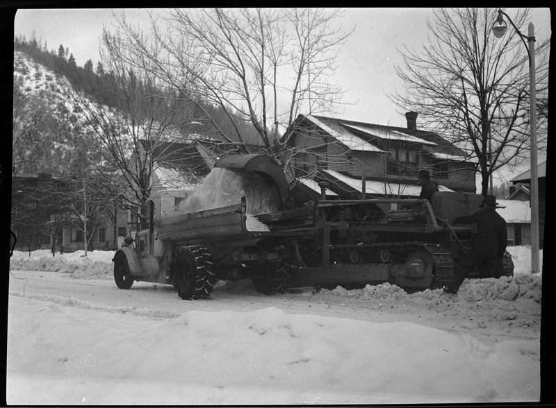 Photo of the back side of a snow loading truck which two men are operating. The truck is actively picking up snow off the street.