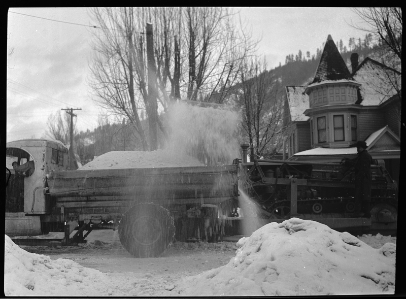 Photo of the side of a snow loading truck in Wallace, Idaho that is actively picking up snow from the street. There is a man at the back of the truck operating it.