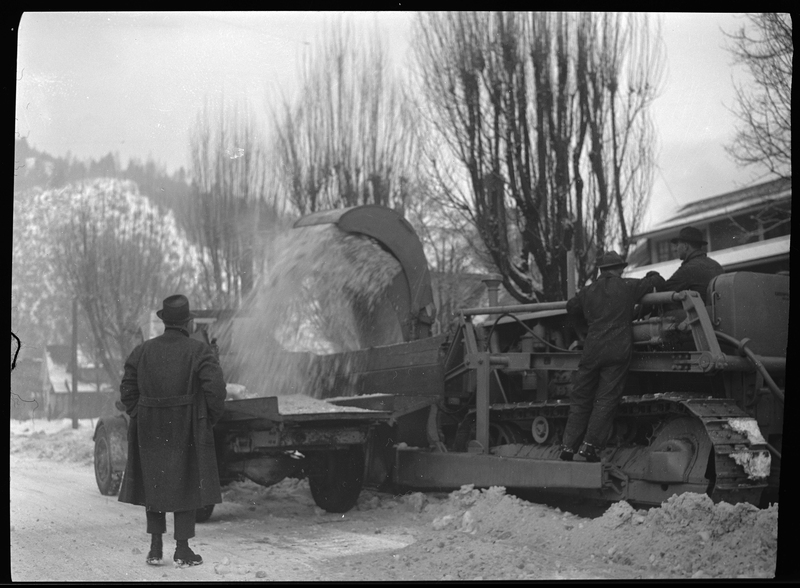 Photo of three men operating the snow loading truck in Wallace, Idaho. The truck is actively removing snow from the ground and dumping it into the truck. One of the men is standing on the machinery, one sits in the truck, and the other observes.