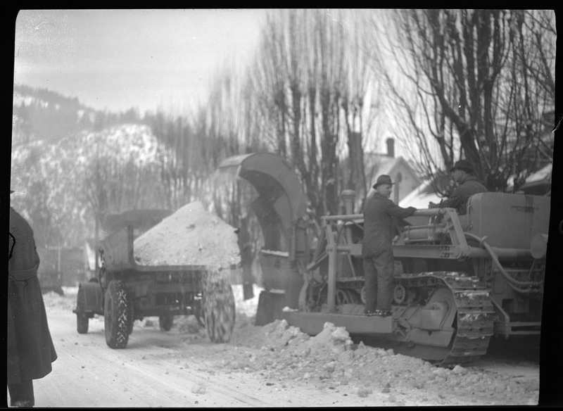 Photo of two men operating the snow loading truck in Wallace, Idaho. The machine is actively picking up and dumping snow into the back of the truck, and the men are operating the machine. One of the men sits in the driver's seat while the other stands on the side.
