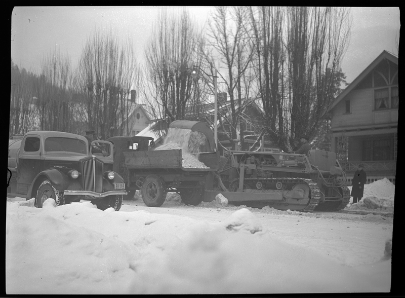 Photo of several men operating the snow loading truck in Wallace, Idaho, which is actively picking up snow from the street and dumping it in the bed of the truck. A car is parked on the other side of the street from the loading truck, and there are several men standing around the equipment to aid in snow removal.