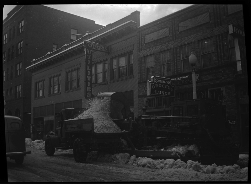 Photo of the back side of the snow loading truck outside the Combination Grocery and Lunch shop in Wallace, Idaho. The machine is actively removing snow from the street.