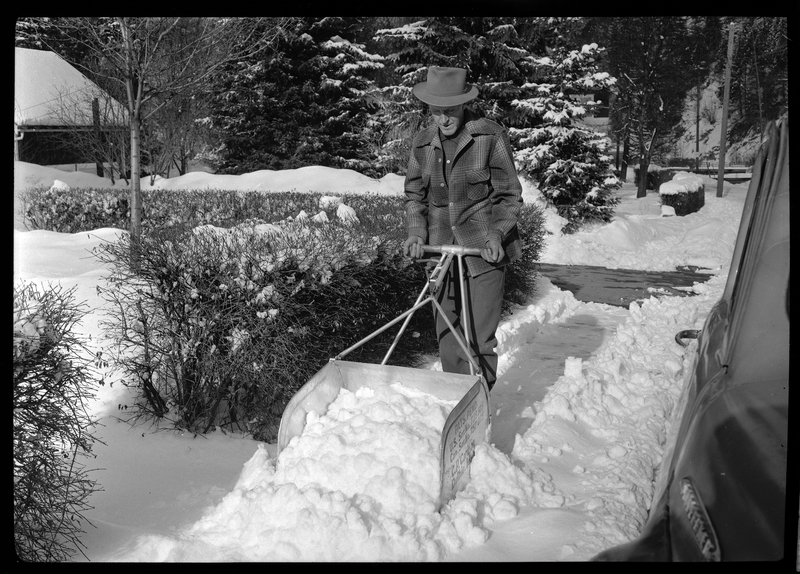 Photo of a man using a C. P. Carlson snow shovel to remove snow from the sidewalk. The shovel requires two hands to push it and holds more snow than the average snow shovel.